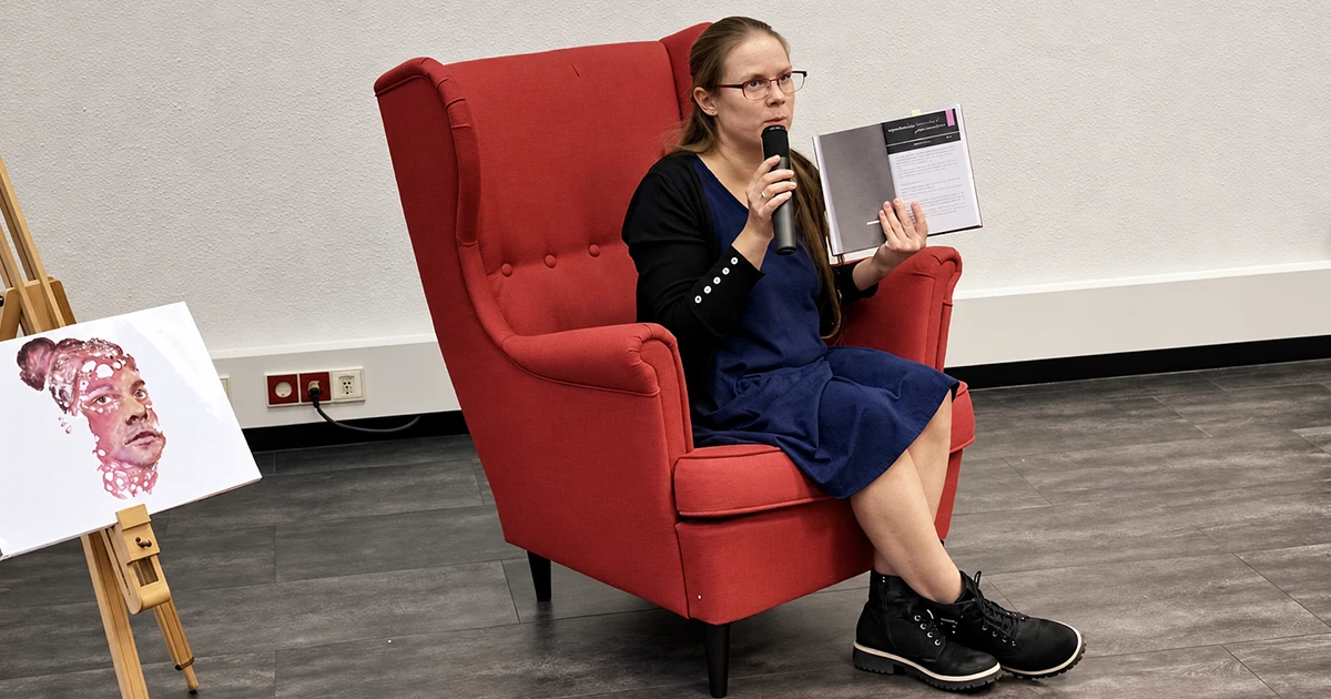 Joke Volkmer sitzt bei ihrer Lesung in Koblenz in einem roten Sessel, hält ihr Buch und spricht in ein Mikrofon.
